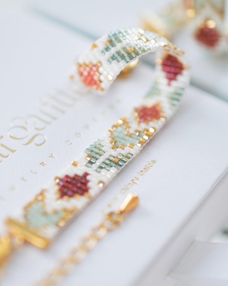 May include: A beaded bracelet with a white background, featuring a repeating pattern of red, green, and gold beads. The bracelet has a gold clasp and chain. The bracelet is on top of a white box.