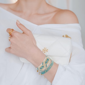 May include: A white quilted leather crossbody bag with a gold chain strap and a gold logo. The image also shows gold rings and two beaded bracelets in turquoise, gold, and white.