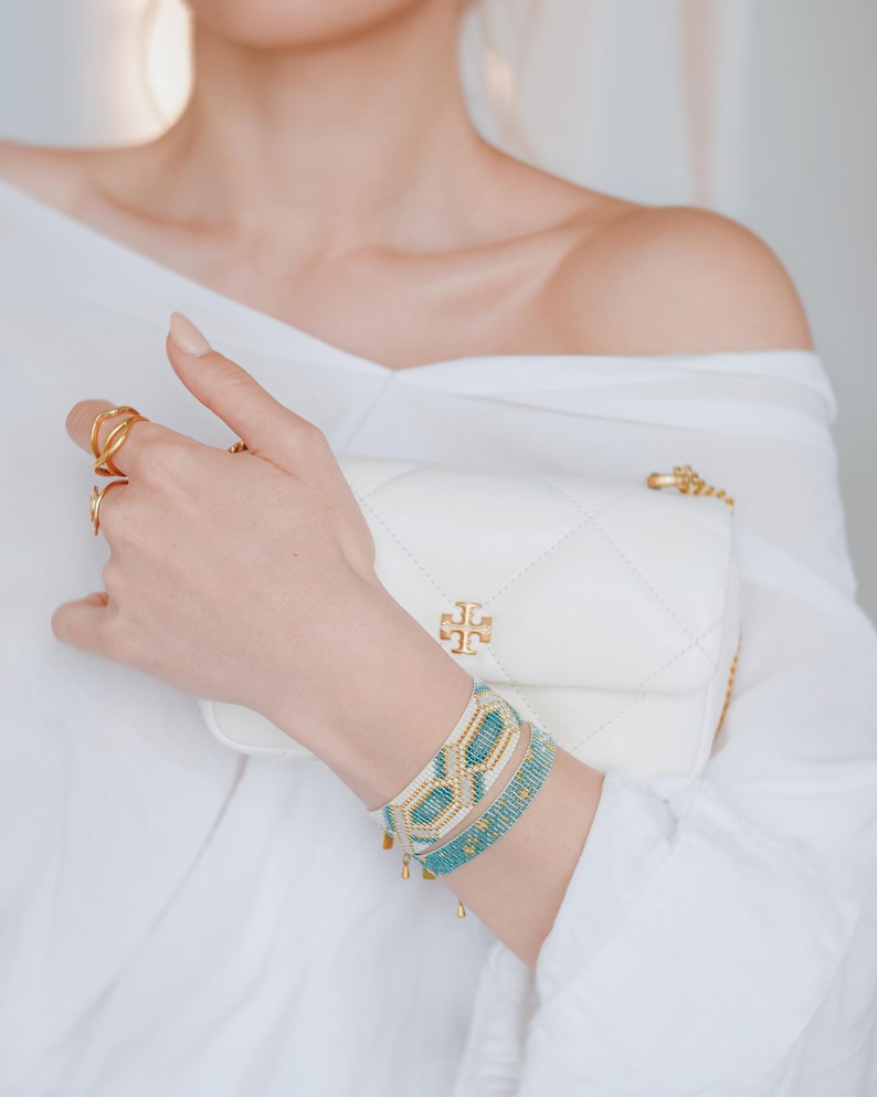 May include: A white quilted leather crossbody bag with a gold chain strap and a gold logo. The image also shows gold rings and two beaded bracelets in turquoise, gold, and white.