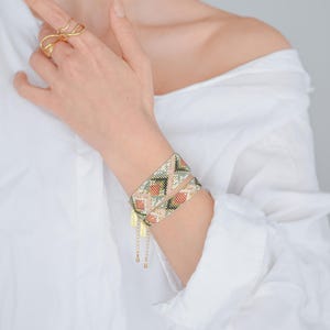 Puede incluir: Dos pulseras envolventes de cuentas con un patr&oacute;n geom&eacute;trico en tonos melocot&oacute;n, verde y dorado se llevan en la mu&ntilde;eca. Las pulseras tienen detalles de cadena y dije dorados. Un anillo dorado se lleva en un dedo.