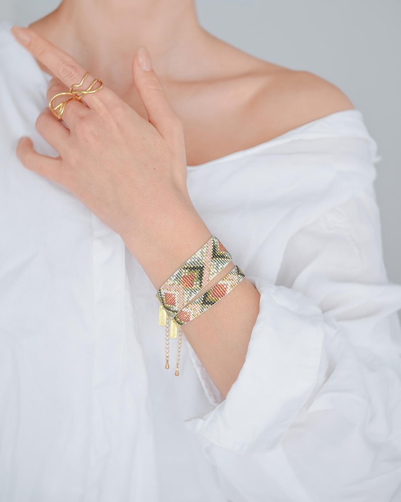 Puede incluir: Dos pulseras envolventes de cuentas con un patr&oacute;n geom&eacute;trico en tonos melocot&oacute;n, verde y dorado se llevan en la mu&ntilde;eca. Las pulseras tienen detalles de cadena y dije dorados. Un anillo dorado se lleva en un dedo.