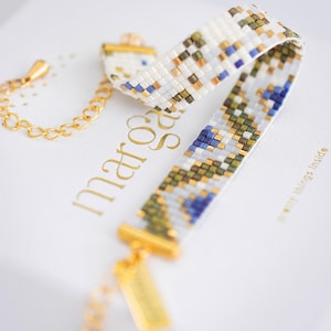 May include: A beaded bracelet with a white base, featuring a pattern of blue, green, and gold beads. The bracelet has gold-colored metal accents and a chain. The bracelet is displayed on a white surface with gold lettering.