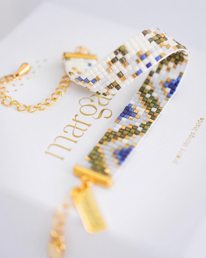 May include: A beaded bracelet with a white base, featuring a pattern of blue, green, and gold beads. The bracelet has gold-colored metal accents and a chain. The bracelet is displayed on a white surface with gold lettering.