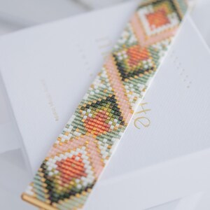 Puede incluir: Una pulsera de cuentas con un patr&oacute;n geom&eacute;trico en tonos rosa, verde, naranja y blanco. La pulsera tiene extremos de metal dorado y un peque&ntilde;o dije. Descansa sobre una superficie blanca.