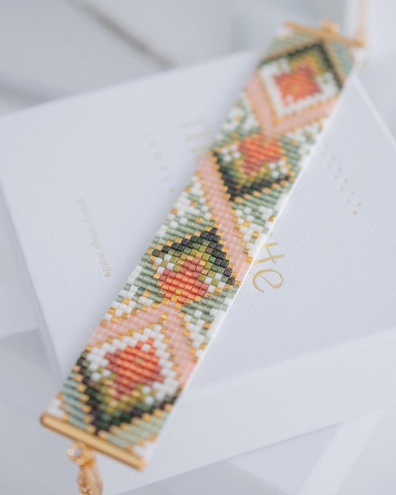 Puede incluir: Una pulsera de cuentas con un patr&oacute;n geom&eacute;trico en tonos rosa, verde, naranja y blanco. La pulsera tiene extremos de metal dorado y un peque&ntilde;o dije. Descansa sobre una superficie blanca.