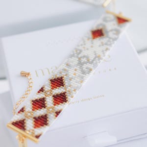 Puede incluir: Un brazalete de cuentas con un patrón blanco, rojo y dorado, apoyado sobre una caja blanca. El brazalete presenta un diseño de corazón repetido en rojo y dorado, con acentos blancos y grises. La caja tiene la palabra "ma" visible.