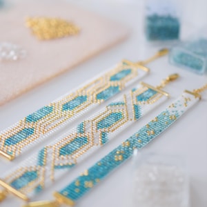 May include: Three beaded bracelets in white, turquoise, and gold. The bracelets have geometric patterns and gold-colored clasps. Beads and crafting supplies are in the background.