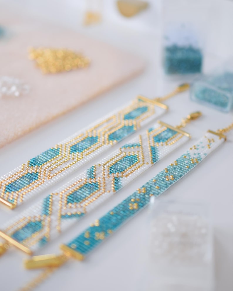 May include: Three beaded bracelets in white, turquoise, and gold. The bracelets have geometric patterns and gold-colored clasps. Beads and crafting supplies are in the background.