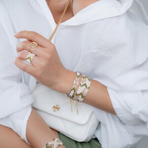 Op de afbeelding: Armbanden met geometrische patronen in groen, roze en wit. Een gouden ring en een witte gewatteerde handtas met een gouden ketting zijn ook zichtbaar. De persoon draagt een wit overhemd.