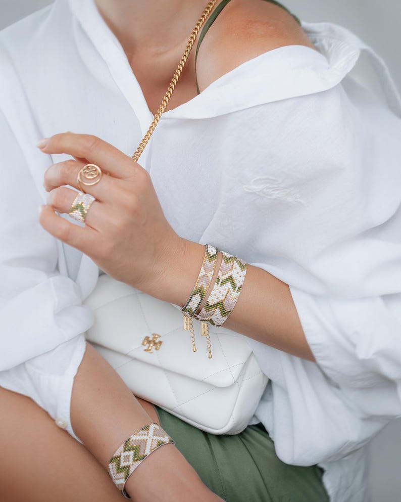 Op de afbeelding: Armbanden met geometrische patronen in groen, roze en wit. Een gouden ring en een witte gewatteerde handtas met een gouden ketting zijn ook zichtbaar. De persoon draagt een wit overhemd.