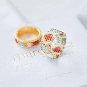 May include: Two beaded rings. One ring is gold with a red, white, and green beaded design. The other ring is white with a red, gold, and green beaded design. The rings are on a white surface with the text "marsige JEWELRY".