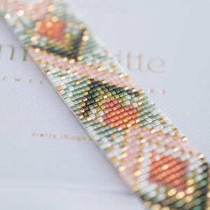 May include: Close-up of a beaded bracelet with a floral pattern. The bracelet features a repeating design of peach, green, and white beads, accented with gold-coloured beads. The bracelet is on a white surface with the text "pretty things".