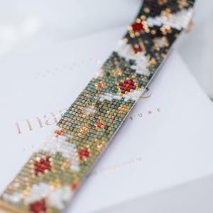 Puede incluir: Una pulsera de cuentas con un cierre dorado. La pulsera presenta un patrón de cuentas rojas, blancas, verdes y doradas. La pulsera descansa sobre una caja blanca con letras doradas. La pulsera mide aproximadamente 2.5 cm de ancho.