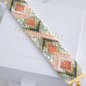 Puede incluir: Una pulsera de cuentas con un patr&oacute;n floral geom&eacute;trico en tonos melocot&oacute;n, verde, blanco y dorado. La pulsera se muestra en una caja blanca con letras doradas. La pulsera mide aproximadamente 2,5 cm de ancho.