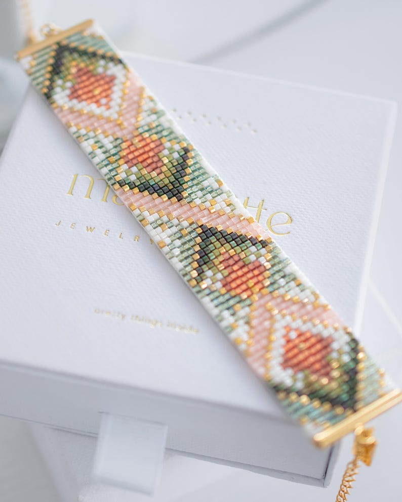 Puede incluir: Una pulsera de cuentas con un patr&oacute;n floral geom&eacute;trico en tonos melocot&oacute;n, verde, blanco y dorado. La pulsera se muestra en una caja blanca con letras doradas. La pulsera mide aproximadamente 2,5 cm de ancho.