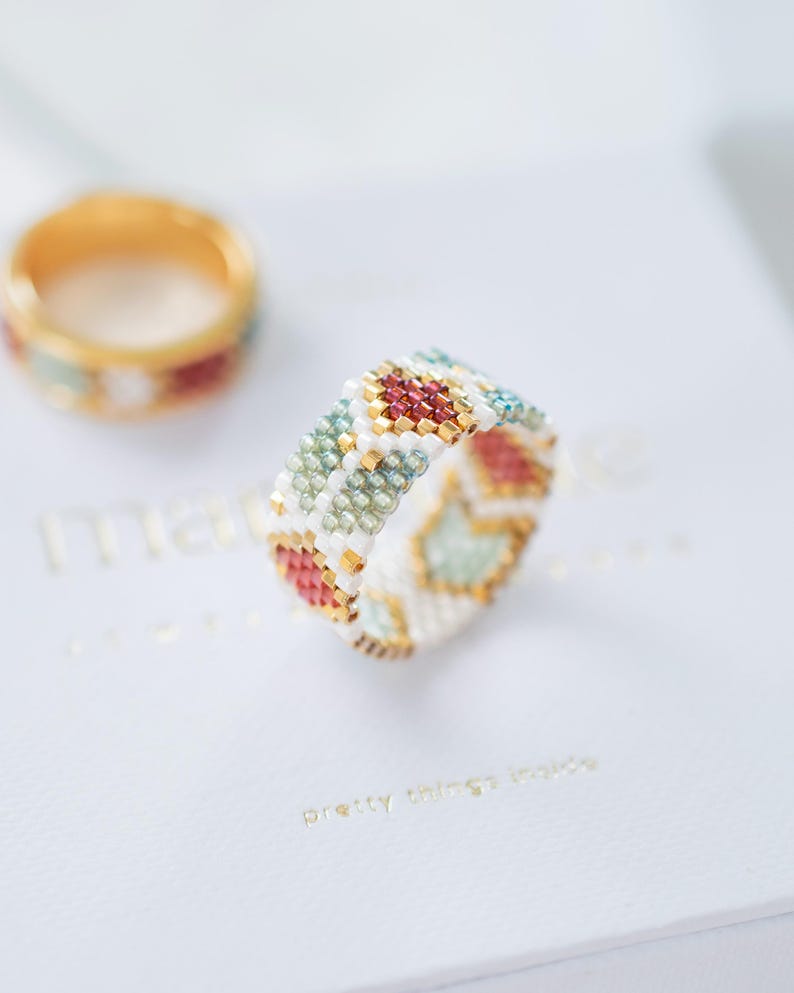 May include: A beaded ring featuring a geometric design in red, gold, white, and teal. Another ring is visible in the background. The rings are set on a white surface with gold text that reads "pretty things inside".