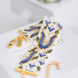 May include: A beaded bracelet with a white background, featuring a floral pattern in blue, green, and gold. The bracelet has gold-colored clasps and a chain. The design is intricate and detailed, showcasing a delicate craft.