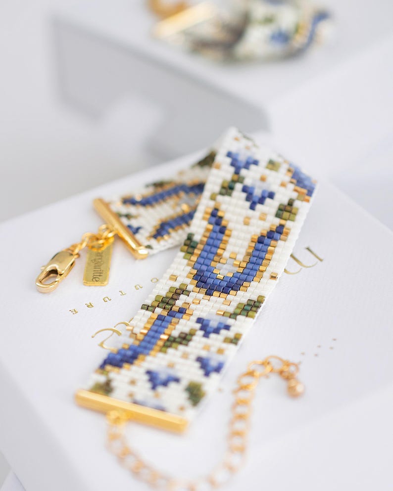 May include: A beaded bracelet with a white background, featuring a floral pattern in blue, green, and gold. The bracelet has gold-colored clasps and a chain. The design is intricate and detailed, showcasing a delicate craft.