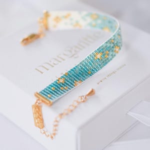 May include: A beaded bracelet with a turquoise and gold pattern, resting on a white box. The bracelet has a gold clasp and chain. The box has the word "Margarita" printed on it.
