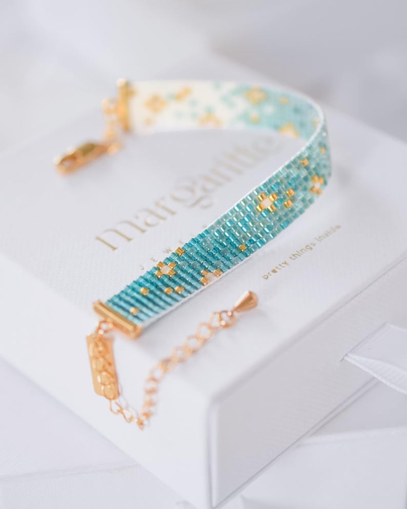 May include: A beaded bracelet with a turquoise and gold pattern, resting on a white box. The bracelet has a gold clasp and chain. The box has the word "Margarita" printed on it.
