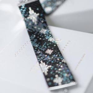Puede incluir: Una pulsera de cuentas con un diseño celestial. La pulsera presenta un fondo negro con patrones de estrellas blancas y turquesas. La pulsera está hecha de pequeñas cuentas cuadradas y tiene un cierre plateado.