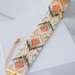 Könnte beinhalten: Ein Armband mit Perlenbesatz und einem floralen Muster in Pfirsich-, Grün- und Weißtönen. Das Armband hat goldfarbene Metallakzente und einen Verschluss. Es liegt auf einer weißen Schachtel.