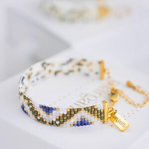 May include: A beaded bracelet with a white base and a geometric pattern in blue, green, and gold. The bracelet has a gold clasp and a small gold tag that says "marguerite".