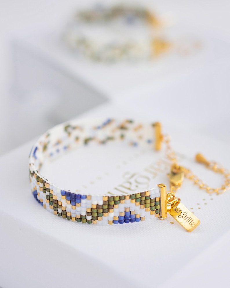 May include: A beaded bracelet with a white base and a geometric pattern in blue, green, and gold. The bracelet has a gold clasp and a small gold tag that says "marguerite".