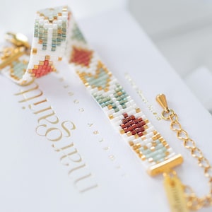 May include: A beaded bracelet with a geometric pattern in white, gold, teal, and rust colors. The bracelet is on a white surface with gold text. The bracelet has a gold clasp and chain.
