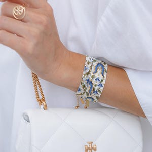 May include: A beaded bracelet with a blue, white, and green floral pattern is worn on a wrist. A gold ring and a white quilted handbag with a gold chain and logo are also visible. The bracelet is approximately 1.5 inches wide.