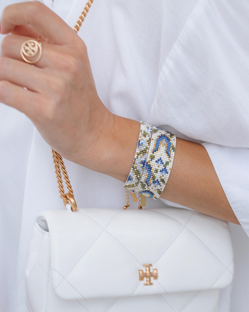 May include: A beaded bracelet with a blue, white, and green floral pattern is worn on a wrist. A gold ring and a white quilted handbag with a gold chain and logo are also visible. The bracelet is approximately 1.5 inches wide.