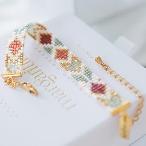 May include: A beaded bracelet with a gold chain and clasp. The bracelet features a geometric pattern in white, gold, pink, and green beads. The bracelet is resting on a white box with gold text.
