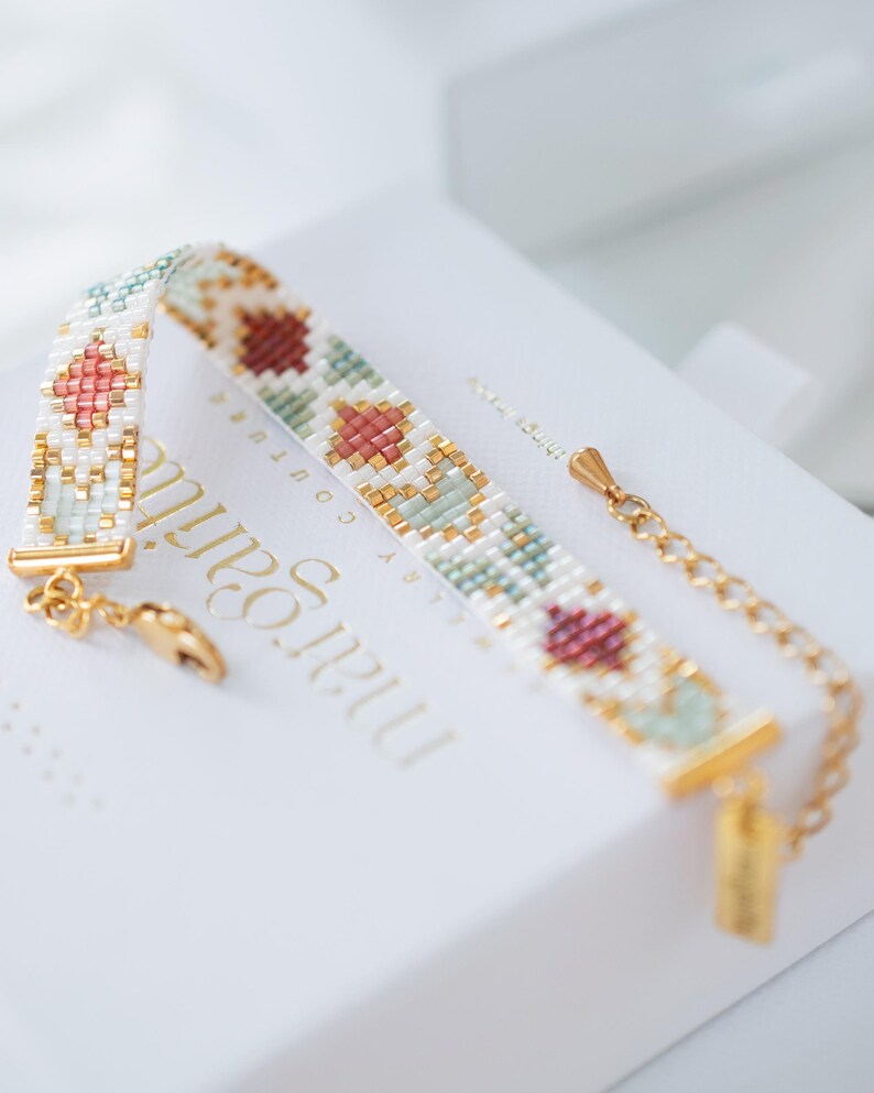 May include: A beaded bracelet with a gold chain and clasp. The bracelet features a geometric pattern in white, gold, pink, and green beads. The bracelet is resting on a white box with gold text.