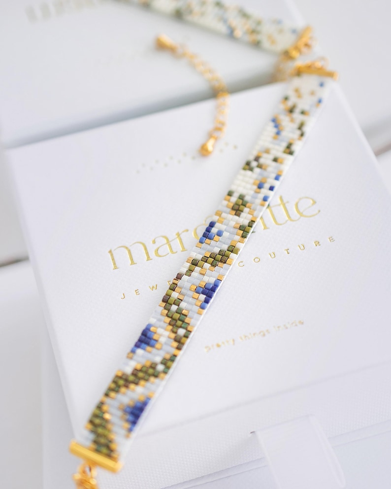 May include: A beaded bracelet with a white base, accented with blue, green, and gold beads. The bracelet is displayed on a white box with gold text that reads "margette JEWELRY COUTURE Pretty things inside."