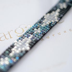 May include: Close-up of a beaded bracelet with a geometric pattern. The bracelet features a mix of iridescent blue, silver, and black beads. The beads are tightly woven together, creating a textured surface. The bracelet is displayed on a white surface.