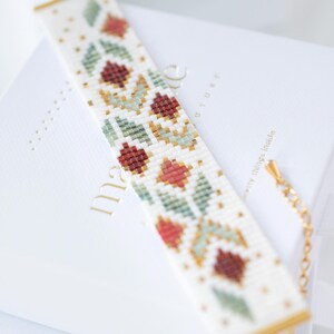 May include: A beaded bracelet with a floral pattern in red, green, and gold on a white background. The bracelet has a gold clasp and accents. The bracelet is approximately 7 inches long.