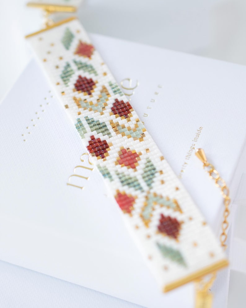 May include: A beaded bracelet with a floral pattern in red, green, and gold on a white background. The bracelet has a gold clasp and accents. The bracelet is approximately 7 inches long.
