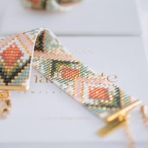 Puede incluir: Una pulsera de cuentas con un patr&oacute;n geom&eacute;trico en tonos melocot&oacute;n, verde, blanco y dorado. La pulsera tiene un cierre y una cadena dorados. La palabra "nate" es visible en la superficie blanca debajo de la pulsera. Joyer&iacute;a.