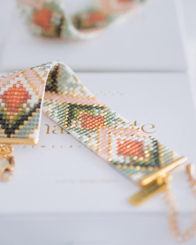 Puede incluir: Una pulsera de cuentas con un patr&oacute;n geom&eacute;trico en tonos melocot&oacute;n, verde, blanco y dorado. La pulsera tiene un cierre y una cadena dorados. La palabra "nate" es visible en la superficie blanca debajo de la pulsera. Joyer&iacute;a.