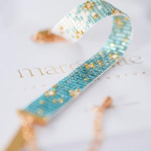 May include: A beaded bracelet with a turquoise and white pattern, accented with gold-colored beads. The bracelet is displayed on a white surface with the brand name "marsaide" in gold lettering. The bracelet has a gold-colored clasp and chain.