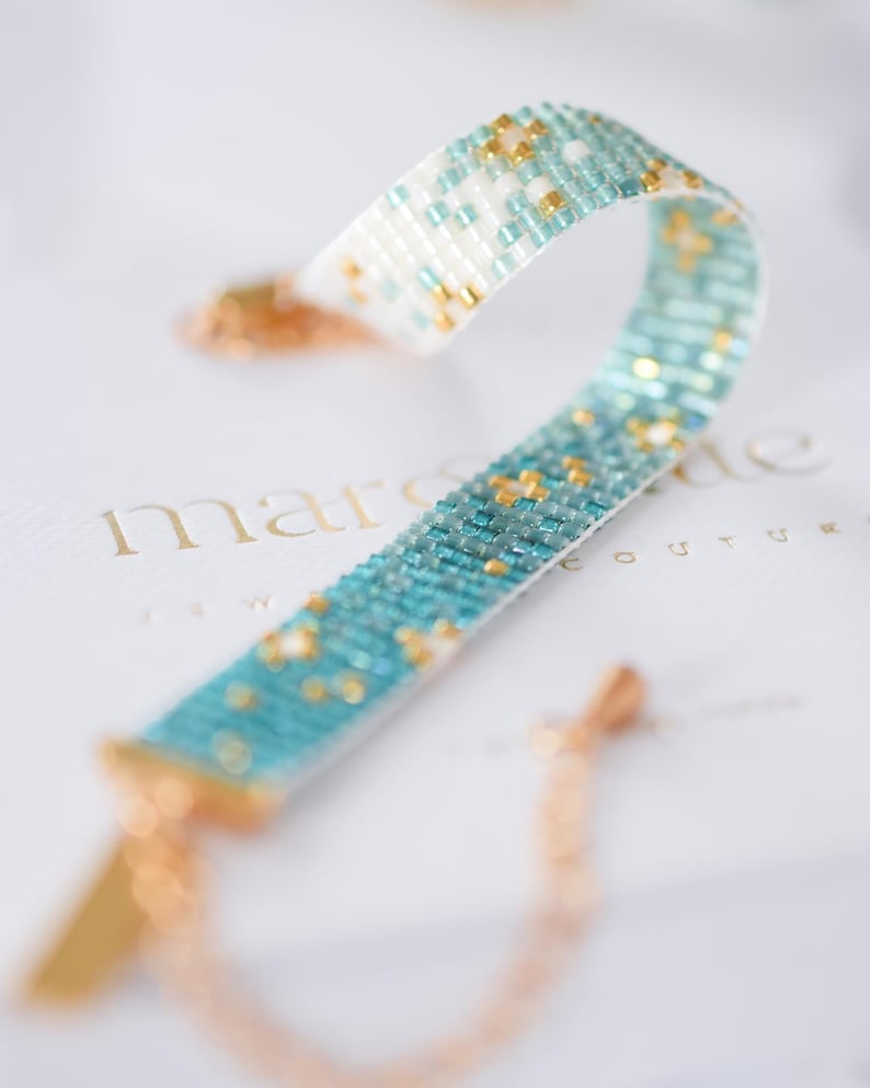 May include: A beaded bracelet with a turquoise and white pattern, accented with gold-colored beads. The bracelet is displayed on a white surface with the brand name "marsaide" in gold lettering. The bracelet has a gold-colored clasp and chain.
