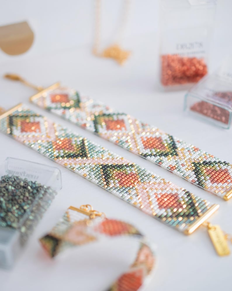 Puede incluir: Dos pulseras de cuentas con un patr&oacute;n geom&eacute;trico en tonos melocot&oacute;n, verde y blanco, con cierres y etiquetas doradas. Las pulseras est&aacute;n dispuestas sobre una superficie blanca, con recipientes de cuentas en el fondo.