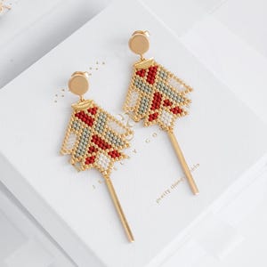 May include: Gold earrings with a geometric design. The earrings have a beaded pattern in red, white, and green, with a gold post and a long, slender gold bar. The earrings are displayed on a white surface.