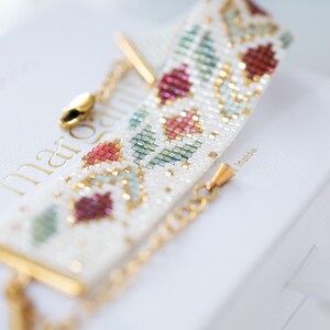 May include: A beaded bracelet with a floral pattern in red, green, and gold on a white background. The bracelet has gold-colored chain and clasp. The bracelet is resting on a white surface.