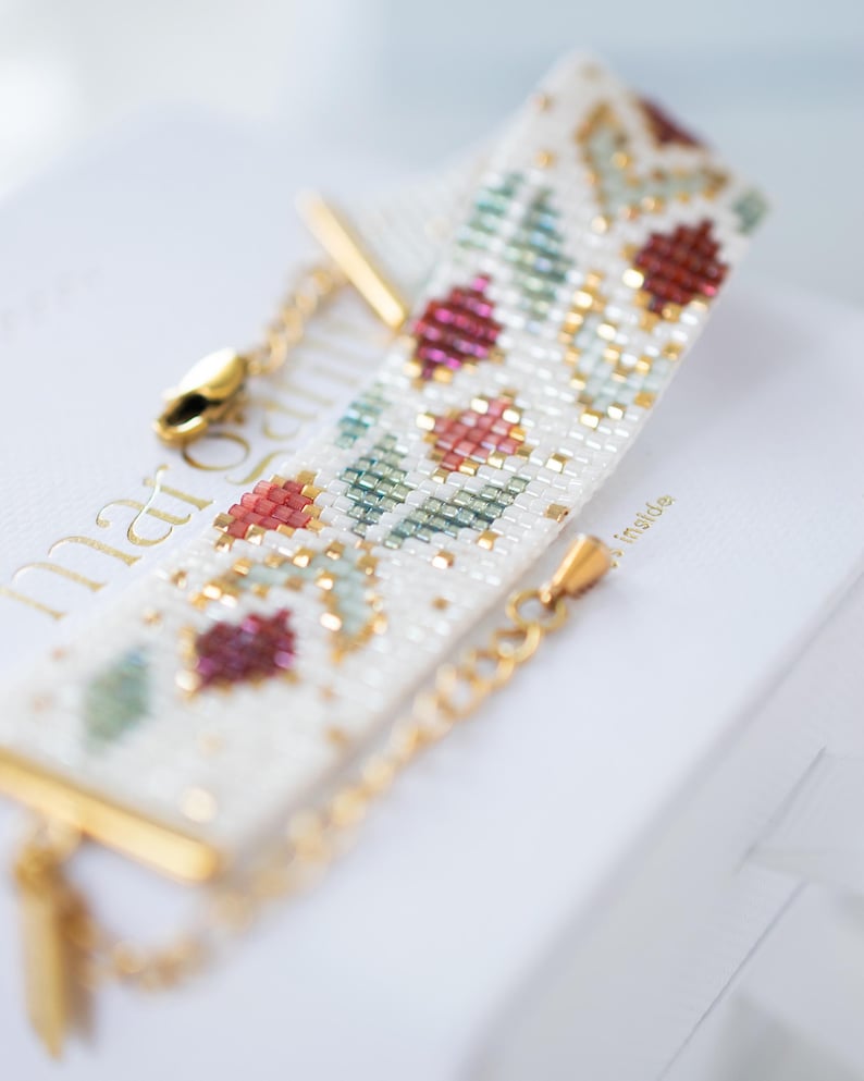 May include: A beaded bracelet with a floral pattern in red, green, and gold on a white background. The bracelet has gold-colored chain and clasp. The bracelet is resting on a white surface.