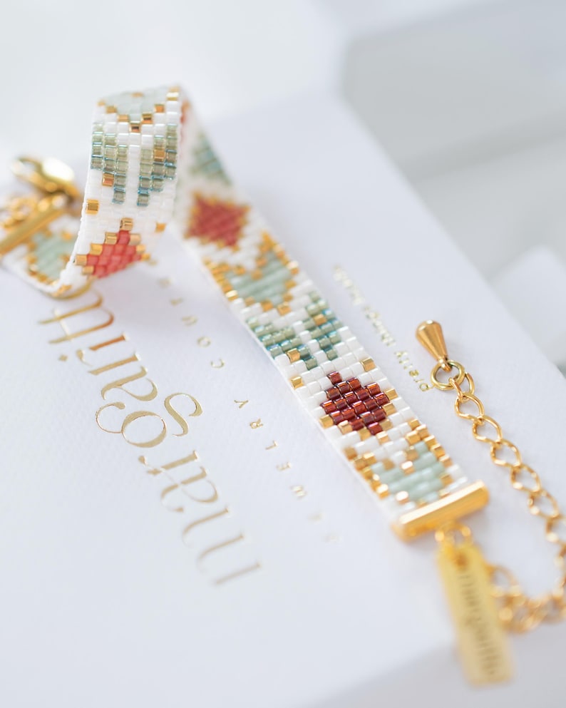 May include: A beaded bracelet with a geometric pattern in white, gold, teal, and rust colors. The bracelet is on a white surface with gold text that reads "margot & co". The bracelet has a gold clasp and chain.