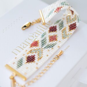 May include: A beaded bracelet with a floral pattern in red, green, and gold on a white background. The bracelet has gold-colored metal accents and a clasp. The bracelet is resting on a white box with gold lettering.