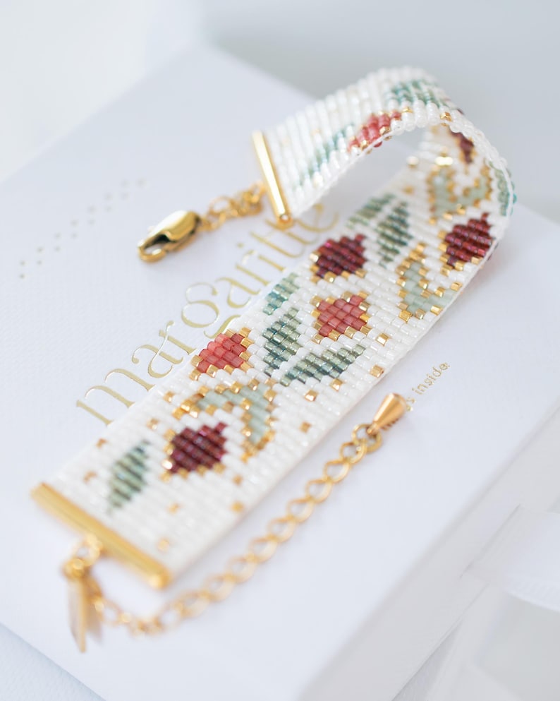 May include: A beaded bracelet with a floral pattern in red, green, and gold on a white background. The bracelet has gold-colored metal accents and a clasp. The bracelet is resting on a white box with gold lettering.