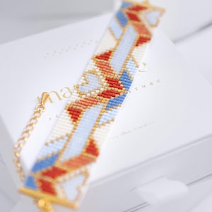 Op de afbeelding: Een kralenarmband met een geometrisch patroon in rood, blauw, wit en goud. De armband ligt op een witte doos met gouden tekst en een gouden ketting sluiting. Het ontwerp van de armband heeft een herhalend patroon van vormen.