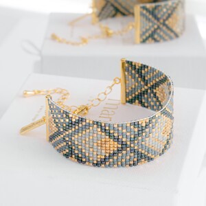 Op de afbeelding: Een kralenarmband met een geometrisch patroon in tinten grijs, goud en blauwgroen. De armband heeft goudkleurige metalen accenten en een kettingsluiting. Een andere armband is zichtbaar op de achtergrond.
